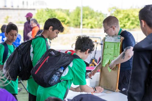 on the left there are two teenagers in green tshirts with backpacks on, leaning over a table and writing on post it notes. On the right is a teacher holding up a board that has lots of yellow post it notes stuck to it. 